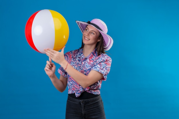 Vrolijke jonge vrouw die met hoed opblaasbare bal houdt die met gelukkig gezicht glimlacht dat zich op blauw bevindt