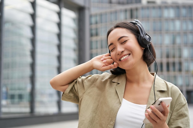 Vrolijke jonge vrouw die op straat danst en muziek luistert in een koptelefoon met een smartphone