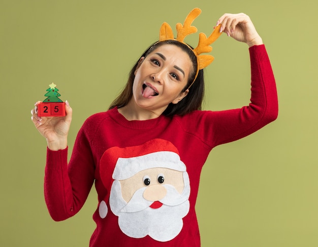 Vrolijke jonge vrouw in rode kerstsweater met grappige rand met hertenhoorns die speelgoedblokjes tonen met datum vijfentwintig die tong uitsteekt die over groene muur staat