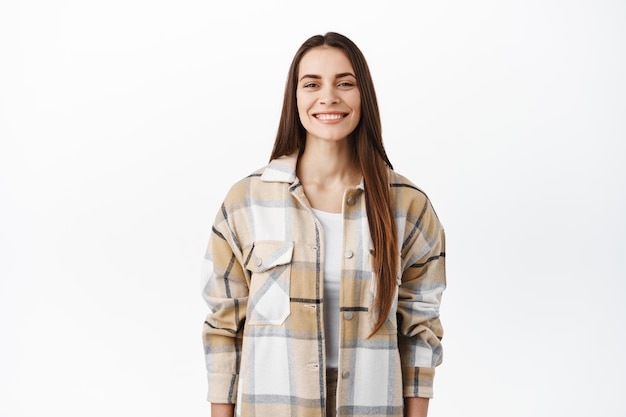 Vrolijke jonge vrouw in stijlvol oversized shirt glimlachend, witte perfecte glimlach tonend, gelukkig kijkend naar de camera, staande op een witte achtergrond