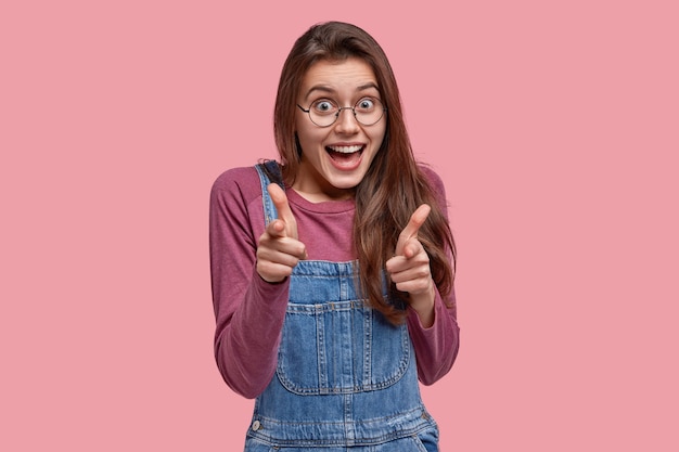 Vrolijke jonge vrouw maakt vinger pistool gebaar op camera, blij om een keuze te maken, draagt denim overall, optische bril