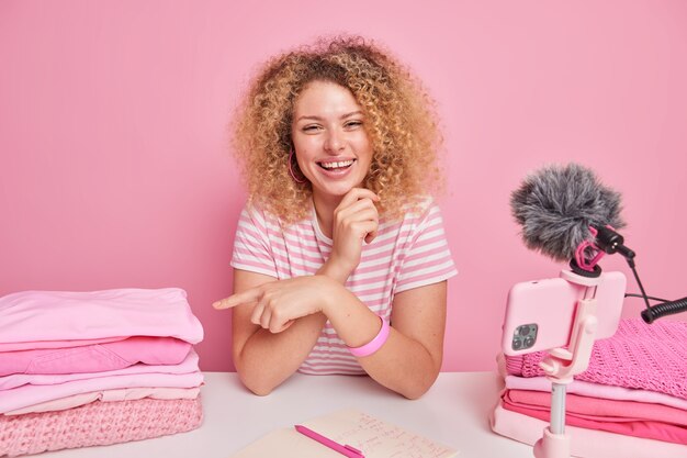 Vrolijke jonge vrouw met krullend haar wijst op stapel schone opgevouwen wasgoed geeft tips over het huishouden voor haar volgelingen maakt aantekeningen in Kladblok zit aan tafel voor smartphonecamera