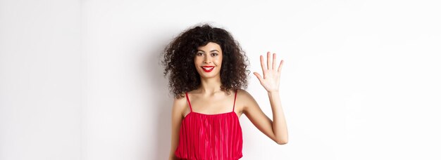 Gratis foto vrolijke jonge vrouw met make-up en rode jurk die vijf vingers toont en glimlachend over wit staat