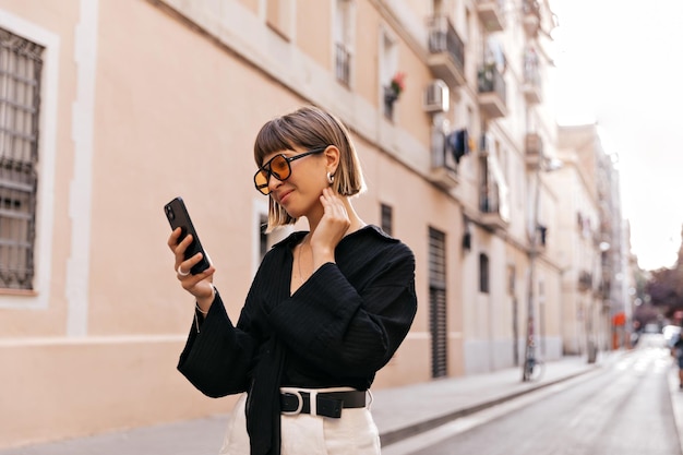 Vrolijke jonge vrouw met mooie glimlach met grijze smartphone in de hand student zakelijke dame Outdoor portret op de achtergrond van de stad Stijlvolle bril zwarte jas dragen