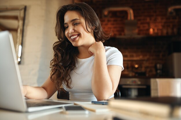 Vrolijke jonge vrouw met pauze tijdens het werken vanuit huis, zittend aan een bureau voor open laptop