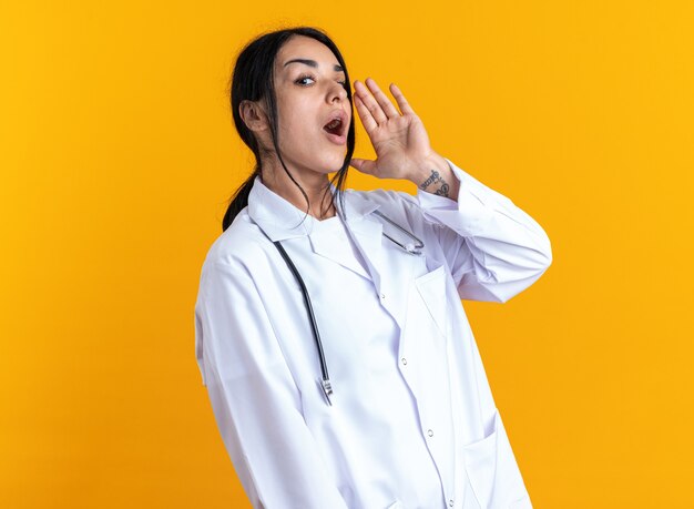 Vrolijke jonge vrouwelijke arts die een medisch gewaad draagt met een stethoscoop die iemand belt die op een gele muur is geïsoleerd