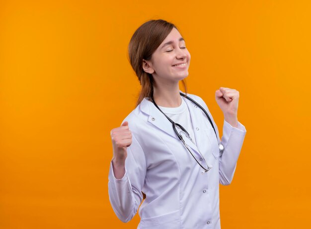 Vrolijke jonge vrouwelijke arts die medische mantel en stethoscoop met gebalde vuisten en gesloten ogen op geïsoleerde oranje ruimte met exemplaarruimte draagt