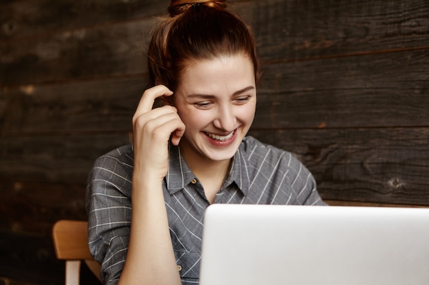 Gratis foto vrolijke jonge vrouwelijke freelancer die voordelen van gratis wi-fi terwijl het werken op afstand op laptop, zittend in coffeeshop. gelukkig student vrouw kijken serie online via internet op notebook pc