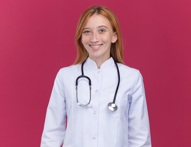 Vrolijke jonge vrouwelijke gemberdokter die een medische mantel en een stethoscoop draagt die lacht