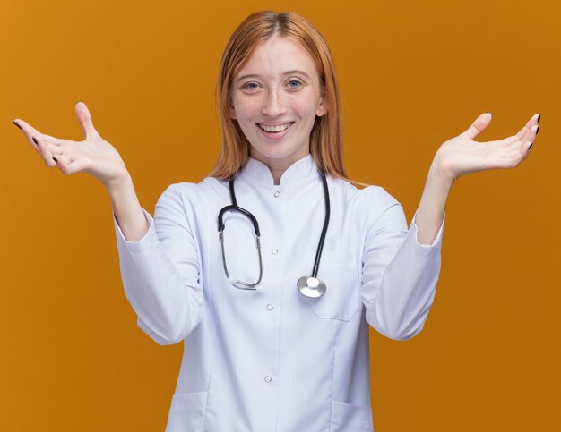 Vrolijke jonge vrouwelijke gemberdokter met een medisch gewaad en een stethoscoop met lege handen geïsoleerd op een oranje muur