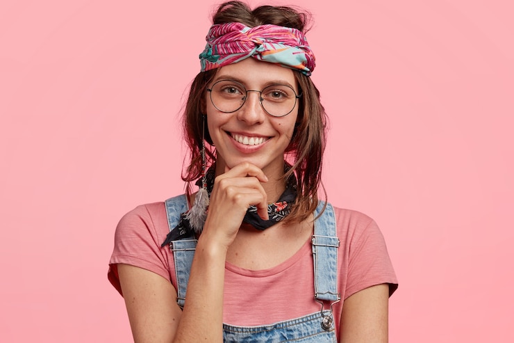 Gratis Foto vrolijke jonge vrouwelijke hippie, houdt kin vast, heeft brede glimlach, draagt modieuze hoofdband en vrijetijdskleding, behoort tot een speciale subcultuur, geïsoleerd over roze muur. mensen en emoties concept