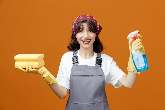 Vrolijke jonge vrouwelijke reiniger met uniforme rubberen handschoenen en bandana met spons en reinigingsmiddel kijkend naar camera geïsoleerd op oranje achtergrond