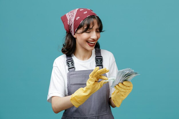Vrolijke jonge vrouwelijke schoonmaker met uniforme bandana en rubberen handschoenen die geld aanhouden en ernaar kijken, geld tellen geïsoleerd op blauwe achtergrond