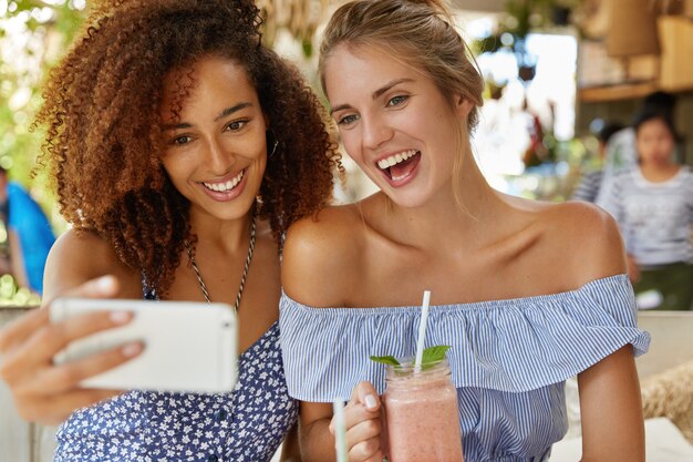 Vrolijke jonge vrouwen of vrouwelijke student die graag poseren voor selfie of videogesprek voeren op slimme telefoon, vrije tijd doorbrengen na lezingen in coffeeshop. Ontspannen lesbisch stel heeft een goed resort in de zomer