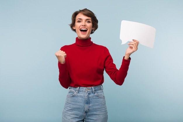 Vrolijke jongedame met donker kort haar in een rode trui die vrolijk ja-gebaar laat zien terwijl ze de witte papieren vorm van het bericht in de hand houdt op een blauwe achtergrond