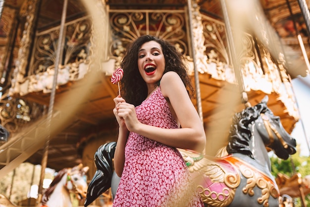 Vrolijke jongedame met donker krullend haar in jurk permanent met lolly pop snoep in handen en gelukkig in de camera kijken met mooie carrousel op achtergrond