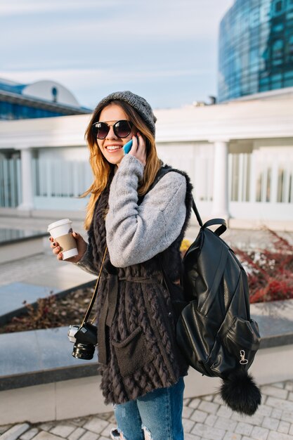 Vrolijke jongedame met koffie gaan wandelen op zonnige koude dag in de grote stad. Mooie vrouw draagt warme winter wollen trui, moderne zonnebril, spreken aan de telefoon, trvelling met camera en tas, gelukkig.