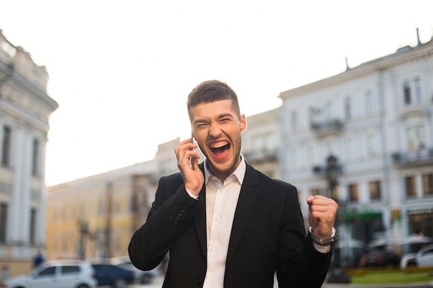 Vrolijke jongeman in zwarte jas en wit overhemd die vrolijk schreeuwt en ja-gebaar toont terwijl hij op mobiel praat met uitzicht op de stad op de achtergrond