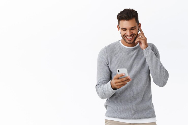 Gratis Foto vrolijke jongeman luistert spraakbericht in smartphone-app met behulp van draadloze oortelefoons die oordopjes aanraken om het volume te verhogen of te verlagen glimlachen en lachen als een mobiel scherm witte achtergrond
