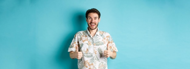 Vrolijke kerel die duimen laat zien op vakantie en reisbureau prijst dat zich in de zomeroverhemd op blauwe b bevindt
