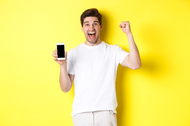Vrolijke kerel die smartphonescherm toont, hand opsteekt en viert, triomfeert over internetprestaties, over gele achtergrond staat