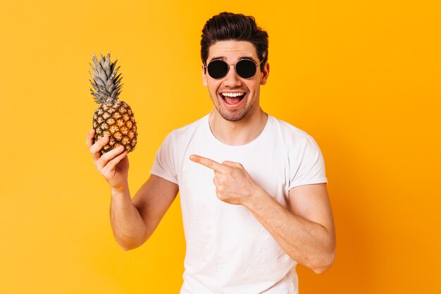 Vrolijke kerel gekleed in wit T-shirt en bril wijst naar ananas en glimlacht op oranje ruimte.