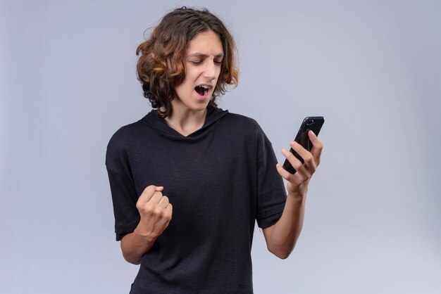 Vrolijke kerel met lang haar in zwart t-shirt met een telefoon op een witte muur