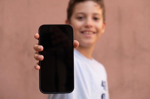 Vrolijke kleine jongen met slimme mobiele telefoon met lege zwarte display. wazig gezicht op de achtergrond geïsoleerd op een lege muur. kinderen die mobiel app-ontwerp tonen voor het inhoudsconcept van kinderen.