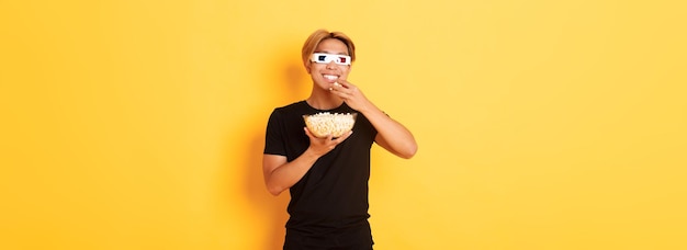 Gratis foto vrolijke knappe aziatische man met blond haar die naar film of tv-series kijkt in een d-bril die popcorn eet en