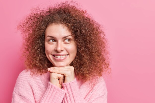 Vrolijke knappe Europese vrouw met bruin krullend houdt handen onder kin glimlacht zachtjes heeft dromerige uitdrukking draagt trui met lange mouwen geïsoleerd over roze muur lege kopie ruimte opzij