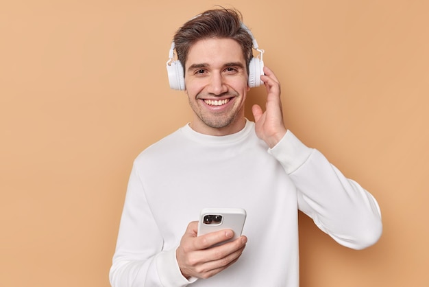 Vrolijke knappe hipster man met brede glimlach bladert smartphone luistert muziek via koptelefoon geniet van goede geluidskwaliteit heeft vrije tijd draagt casual witte trui geïsoleerd over beige achtergrond.