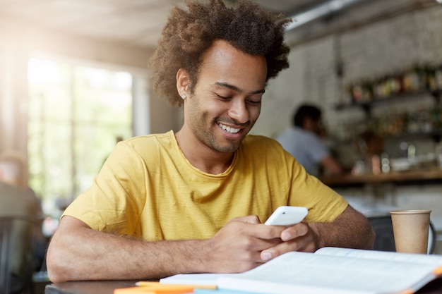 Vrolijke knappe jonge Afro-Amerikaanse mannelijke student in geel t-shirt surfen op internet op slimme telefoon, rust bij koffieshop