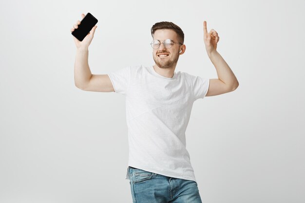 Vrolijke knappe jongen in glazen dansen op muziek in draadloze koptelefoon met mobiele telefoon in de hand