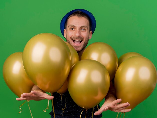 Vrolijke knappe man met blauwe feestmuts staat met helium ballonnen handen geïsoleerd op groene muur met kopie ruimte uitrekken