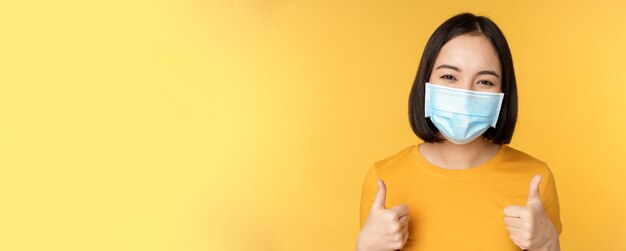 Vrolijke Koreaanse vrouw met medisch gezichtsmasker ondersteunt mensen tijdens pandemie en draagt persoonlijke beschermingsmiddelen van covid19 met duimen omhoog in goedkeuring gele achtergrond