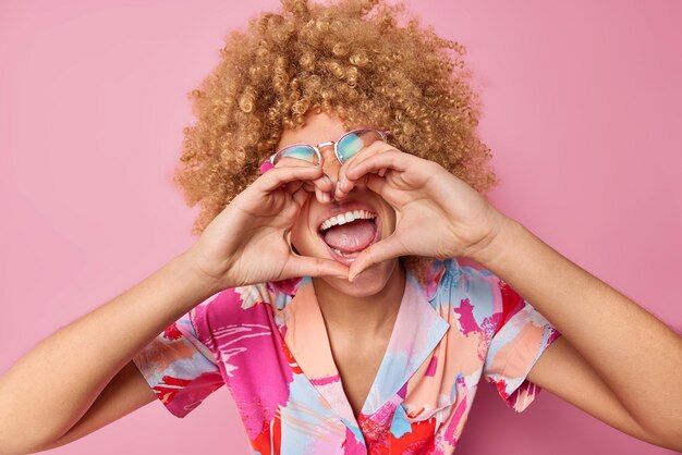 Vrolijke krullend haired vrouw maakt hart gebaar over mond lacht gelukkig liefde uit aan iemands bril en kleurrijke t-shirt geïsoleerd over roze achtergrond romantische mensen concept