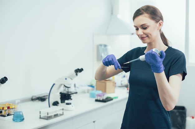 Vrolijke laboratoriumassistent in rubberen handschoenen die een microscoopglaasje vasthoudt en een automatische pipetdispenser gebruikt