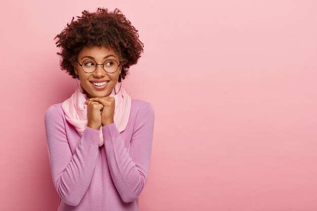 Vrolijke lachende schattige vrouw houdt beide handen onder de kin, kijkt opzij, gekleed in vrijetijdskleding, draagt een bril, geïsoleerd op roze studio achtergrond