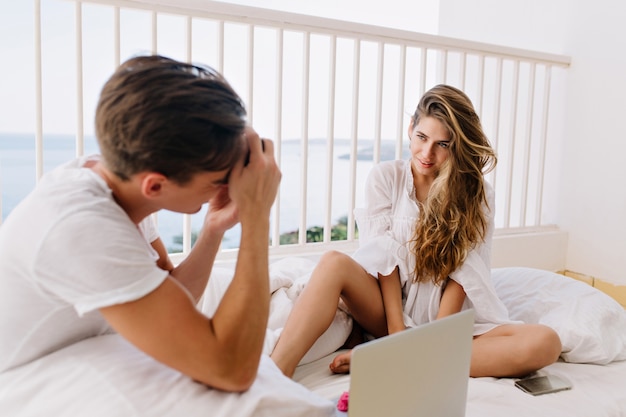 Vrolijke langharige meisje met een gebruinde huid zittend op de witte kussens met telefoon en laptop terwijl haar vriendje foto maakt. man met donker haar nemen foto van zijn geweldige vrouw op balkon