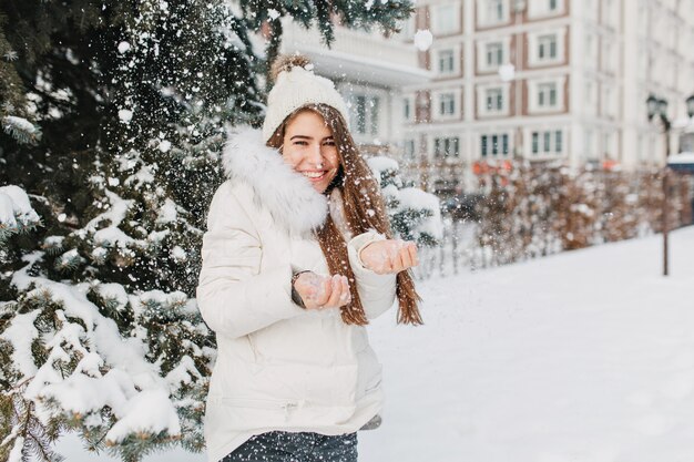 Vrolijke leuke vrouw met plezier met sneeuwvlokken buiten op fir tree vol met sneeuw. Jonge charmante model in warme winterkleren genieten van koud sneeuwt op straat. Positiviteit uiten, glimlachend.