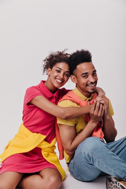 Vrolijke man en zijn vriendin knuffelen op witte achtergrond Optimistisch krullend vrouw in rode jurk en brunette man in gele tee glimlach wijd op geïsoleerde