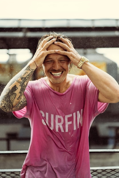 Vrolijke man in een fel t-shirt met een skateboard in een skatepark lacht, water spat uit zijn haar.