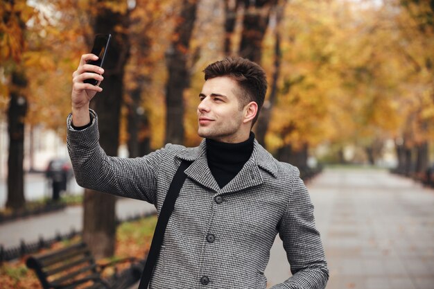 Vrolijke man in jas foto van de natuur nemen of selfie maken met behulp van zwarte smartphone, tijdens het wandelen langs de boulevard