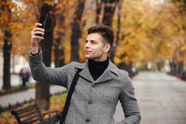 Vrolijke man in jas foto van de natuur nemen of selfie maken met behulp van zwarte smartphone, tijdens het wandelen langs de boulevard