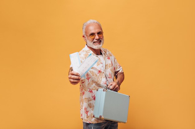 Vrolijke man in plant print shirt poseert met kaartjes en koffer Bebaarde man met een glimlach op zijn gezicht in lichte outfit poseren op oranje achtergrond