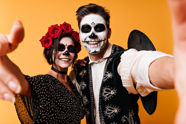 Vrolijke man in sombrero selfie met vriendin maken in halloween. Grappige jongens met zombiemake-up die op oranje muur koelen.