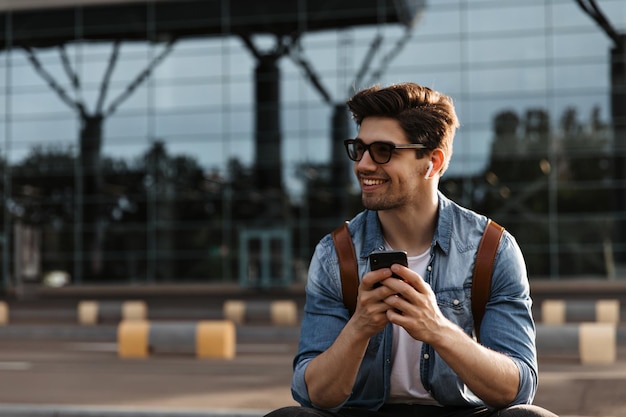 Vrolijke man in zonnebril en spijkerjasje lacht breed. Gelukkige brunette man in witte t-shirt houdt telefoon vast en zit buiten