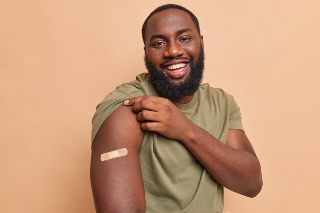 Vrolijke man toont hechtpleister op schouder nadat hij coronavirusvaccin heeft gekregen voelt zich veilig krijgt injectie in arm geeft om gezondheid tijdens pandemie geïsoleerd over beige muur