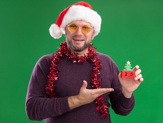 Vrolijke man van middelbare leeftijd met een kerstmuts en een klatergoudslinger om de nek met een bril die vasthoudt en met de hand wijst naar kerstboomspeelgoed met datum die naar camera kijkt geïsoleerd op groene achtergrond