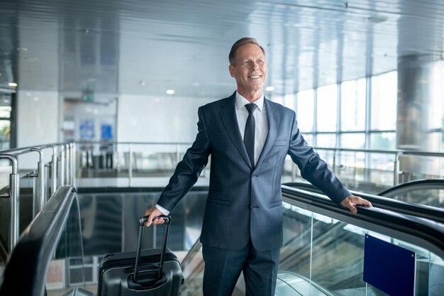 Vrolijke mannelijke passagier met de trolleykoffer die van de roltrap op de luchthaven stapt